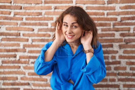 Photo for Beautiful brunette woman standing over bricks wall trying to hear both hands on ear gesture, curious for gossip. hearing problem, deaf - Royalty Free Image