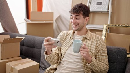 Photo for Young hispanic man using smartphone drinking coffee at new home - Royalty Free Image