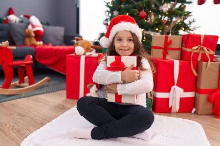 Téléchargez les photos : Adorable fille hispanique étreignant cadeau de Noël assis sur le sol à la maison - en image libre de droit