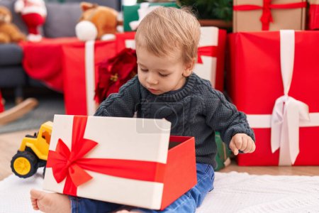 Téléchargez les photos : Adorable tout-petit blond déballer cadeau de Noël assis sur le sol à la maison - en image libre de droit