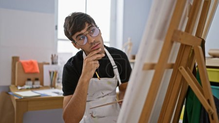 Photo for Serious young hispanic man artist sitting in art studio, thinking and doubting over his drawing on canvas, concentrated in idea amid a cascade of paintbrushes, palette and easel. - Royalty Free Image