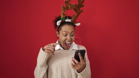 Téléchargez les photos : Femme afro-américaine utilisant un smartphone portant des oreilles de renne sur fond rouge isolé - en image libre de droit