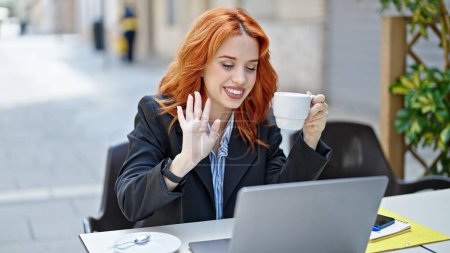 Téléchargez les photos : Jeune rousse femme d'affaires ayant appel vidéo boire du café à la terrasse du café - en image libre de droit