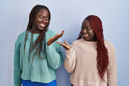 Foto de Dos mujeres africanas de pie sobre fondo azul sonriendo mostrando ambas manos palmas abiertas, presentando y comparando publicidad y equilibrio - Imagen libre de derechos