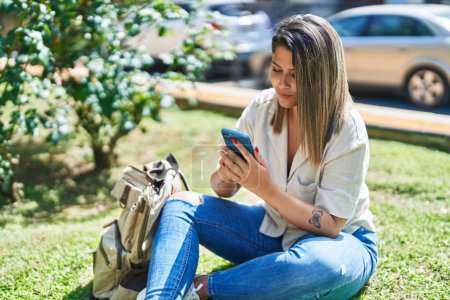 Téléchargez les photos : Jeune étudiante hispanique utilisant un smartphone assis sur l'herbe au parc - en image libre de droit