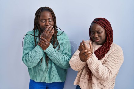 Téléchargez les photos : Deux femmes africaines debout sur fond bleu souffrant de douleurs aux mains et aux doigts, inflammation de l'arthrite - en image libre de droit
