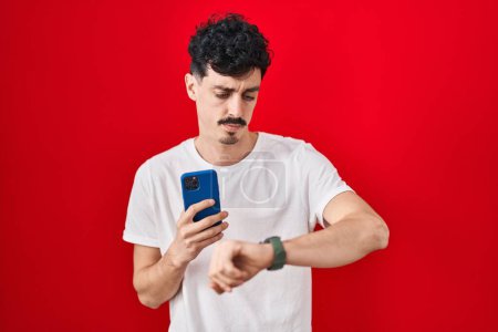 Téléchargez les photos : Homme hispanique en utilisant smartphone sur fond rouge vérification de l'heure sur montre-bracelet, détendu et confiant - en image libre de droit