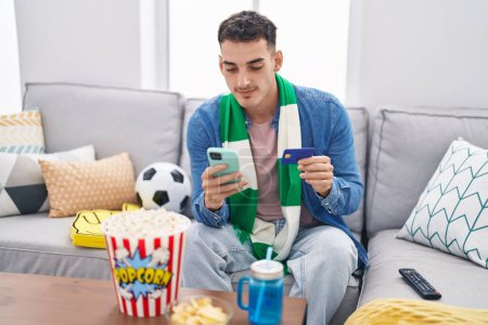 Téléchargez les photos : Jeune homme hispanique fan de football utilisant smartphone et carte de crédit assis sur le canapé à la maison - en image libre de droit