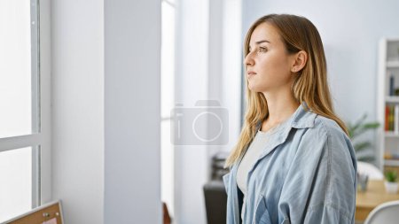 Téléchargez les photos : Jeune femme d'affaires blonde au visage sérieux, regardant intensément par la fenêtre du bureau, montrant sa concentration au travail - en image libre de droit