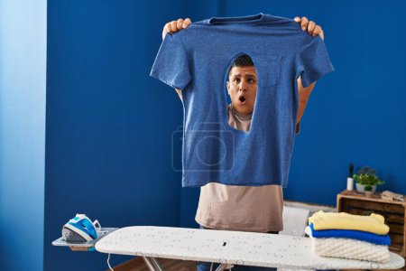 Photo for Young latin man holding t shirt with burned hole with surprise expression at laundry room - Royalty Free Image