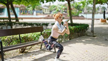 Foto de Mujer pelirroja joven escuchando música entrenamiento piernas ejercicio en el parque - Imagen libre de derechos