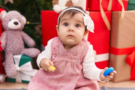 Téléchargez les photos : Adorable bébé hispanique tenant cerceaux jouet assis sur le sol près de l'arbre de Noël à la maison - en image libre de droit