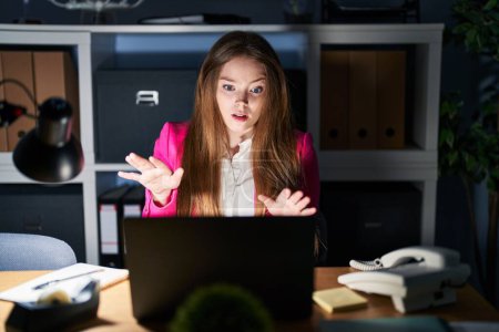 Foto de Young caucasian woman working at the office at night afraid and terrified with fear expression stop gesture with hands, shouting in shock. panic concept. - Imagen libre de derechos