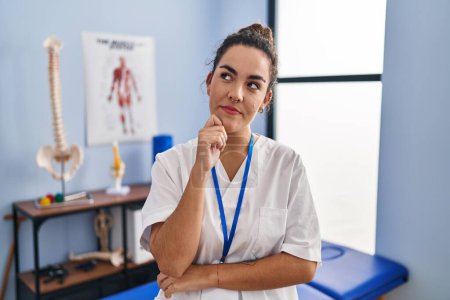 Photo for Young hispanic woman working at rehabilitation clinic with hand on chin thinking about question, pensive expression. smiling with thoughtful face. doubt concept. - Royalty Free Image