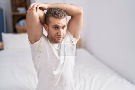 Téléchargez les photos : Jeune homme caucasien étirant le bras assis sur le lit dans la chambre - en image libre de droit