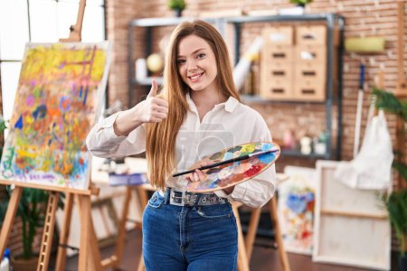 Foto de Joven mujer caucásica en el estudio de arte sosteniendo paleta sonriendo feliz y positivo, pulgar hacia arriba haciendo excelente y signo de aprobación - Imagen libre de derechos