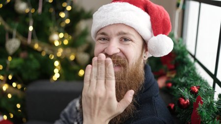 Téléchargez les photos : Jeune rousse homme souriant confiant assis près de l'arbre de Noël avec la main sur la bouche à la maison - en image libre de droit