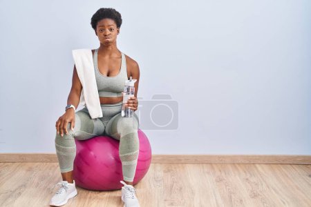 Foto de Mujer afroamericana usando ropa deportiva sentada en pilates bola soplando mejillas con cara divertida. boca hinchada de aire, expresión loca. - Imagen libre de derechos
