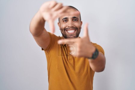 Foto de Hombre hispano con barba de pie sobre fondo blanco sonriendo haciendo marco con las manos y los dedos con la cara feliz. concepto de creatividad y fotografía. - Imagen libre de derechos