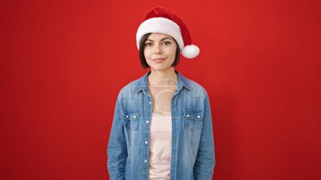 Téléchargez les photos : Jeune femme caucasienne souriante confiante portant un chapeau de Noël sur fond rouge isolé - en image libre de droit