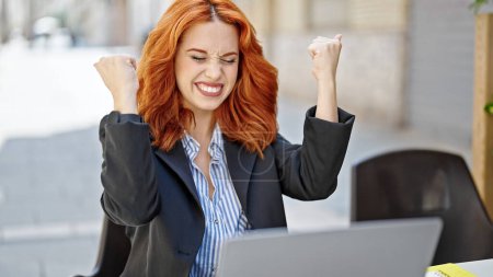 Téléchargez les photos : Jeune rousse femme d'affaires utilisant un ordinateur portable avec expression gagnante sur la terrasse du café - en image libre de droit