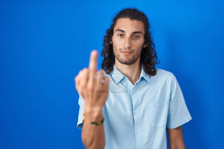 Foto de Young hispanic man standing over blue background showing middle finger, impolite and rude fuck off expression - Imagen libre de derechos