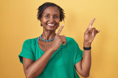 Foto de Mujer africana con rastas de pie sobre fondo amarillo sonriendo y mirando a la cámara apuntando con dos manos y dedos hacia un lado. - Imagen libre de derechos