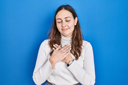 Photo for Young hispanic woman standing over blue background smiling with hands on chest with closed eyes and grateful gesture on face. health concept. - Royalty Free Image