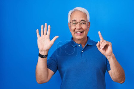 Foto de Hombre de mediana edad con el pelo gris de pie sobre fondo azul mostrando y apuntando hacia arriba con los dedos número seis mientras sonríe confiado y feliz. - Imagen libre de derechos