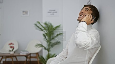 Photo for Confident young hispanic man engages in smiling business conversation, sitting on chair in waiting room, managing work call indoors on smartphone - Royalty Free Image