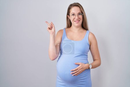 Foto de Mujer embarazada joven de pie sobre fondo blanco con una gran sonrisa en la cara, señalando con el dedo de la mano a un lado mirando a la cámara. - Imagen libre de derechos