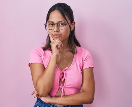 Foto de Mujer joven hispana de pie sobre fondo rosa con gafas que miran con confianza a la cámara con sonrisa con los brazos cruzados y la mano levantada en la barbilla. pensar positivamente. - Imagen libre de derechos