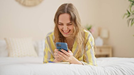 Photo for Young blonde woman using smartphone lying on bed smiling at bedroom - Royalty Free Image