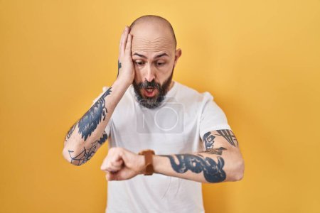 Foto de Joven hombre hispano con tatuajes de pie sobre fondo amarillo mirando la hora del reloj preocupado, con miedo de llegar tarde - Imagen libre de derechos