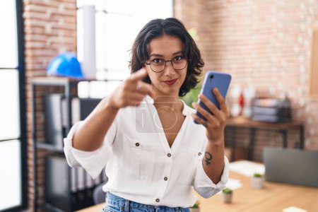 Photo for Young hispanic woman working at the office using smartphone pointing with finger to the camera and to you, confident gesture looking serious - Royalty Free Image