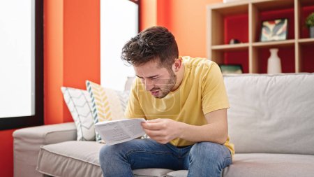 Téléchargez les photos : Jeune homme hispanique lisant le document assis sur le canapé à la maison - en image libre de droit