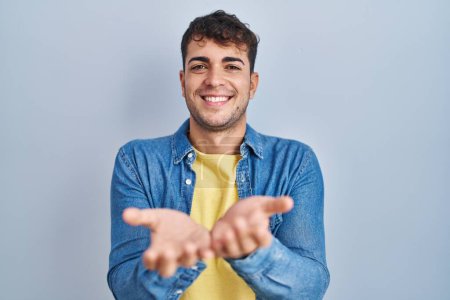 Photo for Young hispanic man standing over blue background smiling with hands palms together receiving or giving gesture. hold and protection - Royalty Free Image