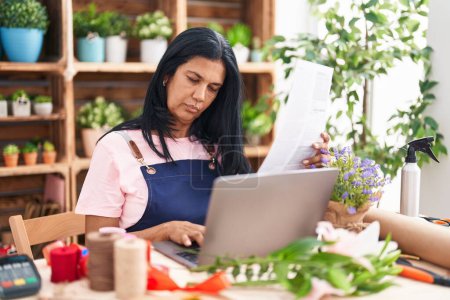 Foto de Middle age hispanic woman florist using laptop reading document at florist - Imagen libre de derechos
