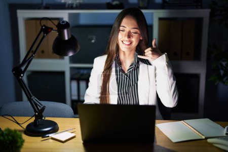 Téléchargez les photos : Jeune femme brune travaillant au bureau la nuit avec un ordinateur portable souriant faisant geste de téléphone avec la main et les doigts comme parler au téléphone. communiquer les concepts. - en image libre de droit