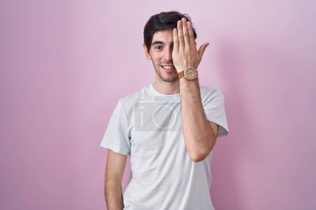 Photo for Young hispanic man standing over pink background covering one eye with hand, confident smile on face and surprise emotion. - Royalty Free Image
