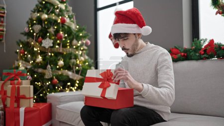 Photo for Young hispanic man wearing christmas hat unpacking gift with surprised face at home - Royalty Free Image
