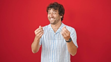 Téléchargez les photos : Jeune homme hispanique souriant confiant faire geste d'argent sur fond rouge isolé - en image libre de droit