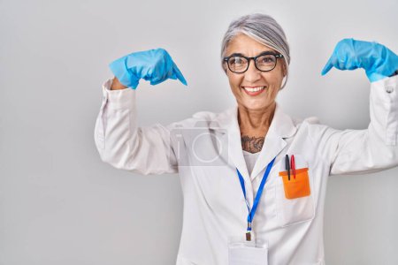 Foto de Mujer de mediana edad con el pelo gris vistiendo túnica científica mirando confiado con sonrisa en la cara, señalándose con los dedos orgullosos y felices. - Imagen libre de derechos