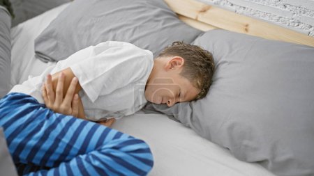 Photo for Adorable blond boy experiencing stomach pain, lying on bed in his cozy bedroom, touching his abdomen, suffering visibly - Royalty Free Image