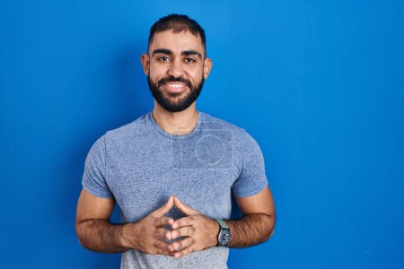 Téléchargez les photos : Moyen-Orient homme avec barbe debout sur fond bleu mains ensemble et doigts croisés souriant détendu et joyeux. succès et optimisme - en image libre de droit