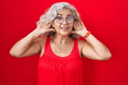 Photo for Middle age woman with grey hair standing over red background trying to hear both hands on ear gesture, curious for gossip. hearing problem, deaf - Royalty Free Image