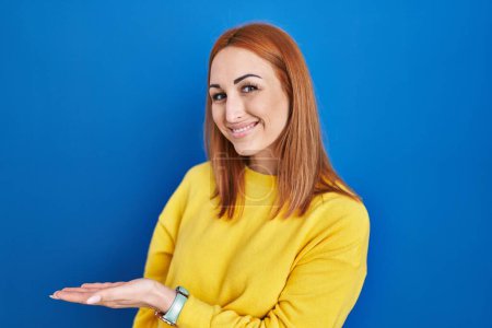 Foto de Young woman standing over blue background inviting to enter smiling natural with open hand - Imagen libre de derechos