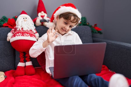 Foto de Adorable chico hispano teniendo videollamada sentado en el sofá por la decoración de Navidad en casa - Imagen libre de derechos