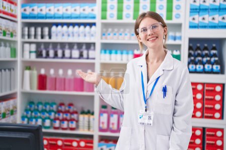 Foto de Joven mujer rubia farmacéutica sonriendo confiado presentando en la farmacia - Imagen libre de derechos