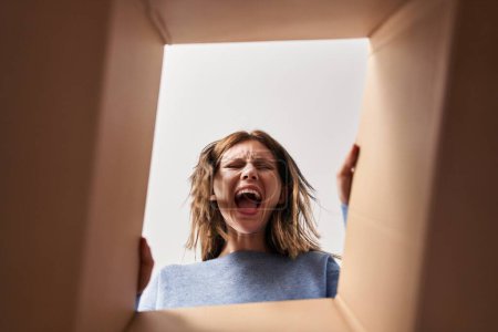 Photo for Beautiful woman opening cardboard box angry and mad screaming frustrated and furious, shouting with anger looking up. - Royalty Free Image